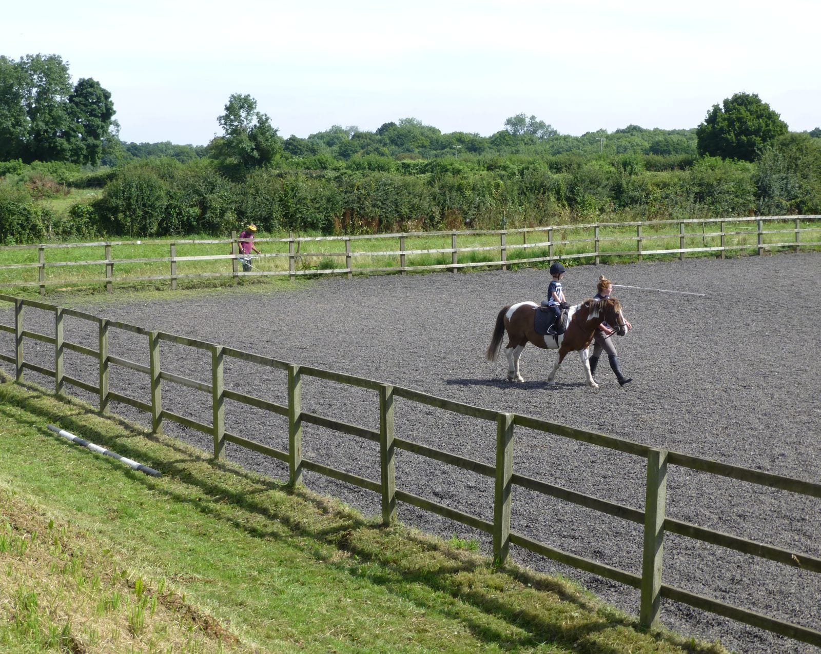 Young child in a riding hat sitting on a pony at walk in a fenced outdoor sand school, led on foot by an instructor, with English countryside beyond the rails