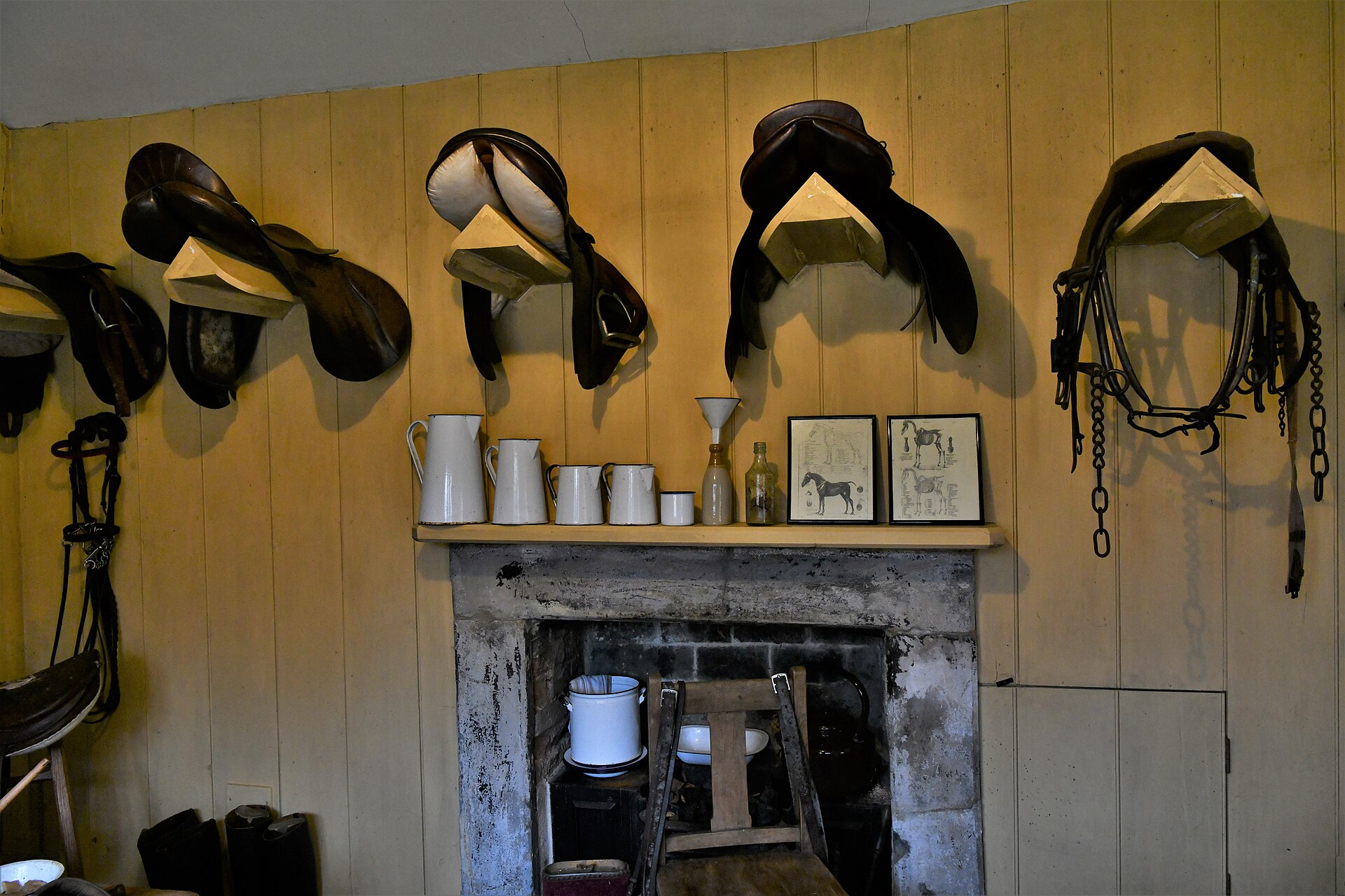 Estate tack room with English saddles on wall racks, bridles on hooks and horse-care prints above the hearth