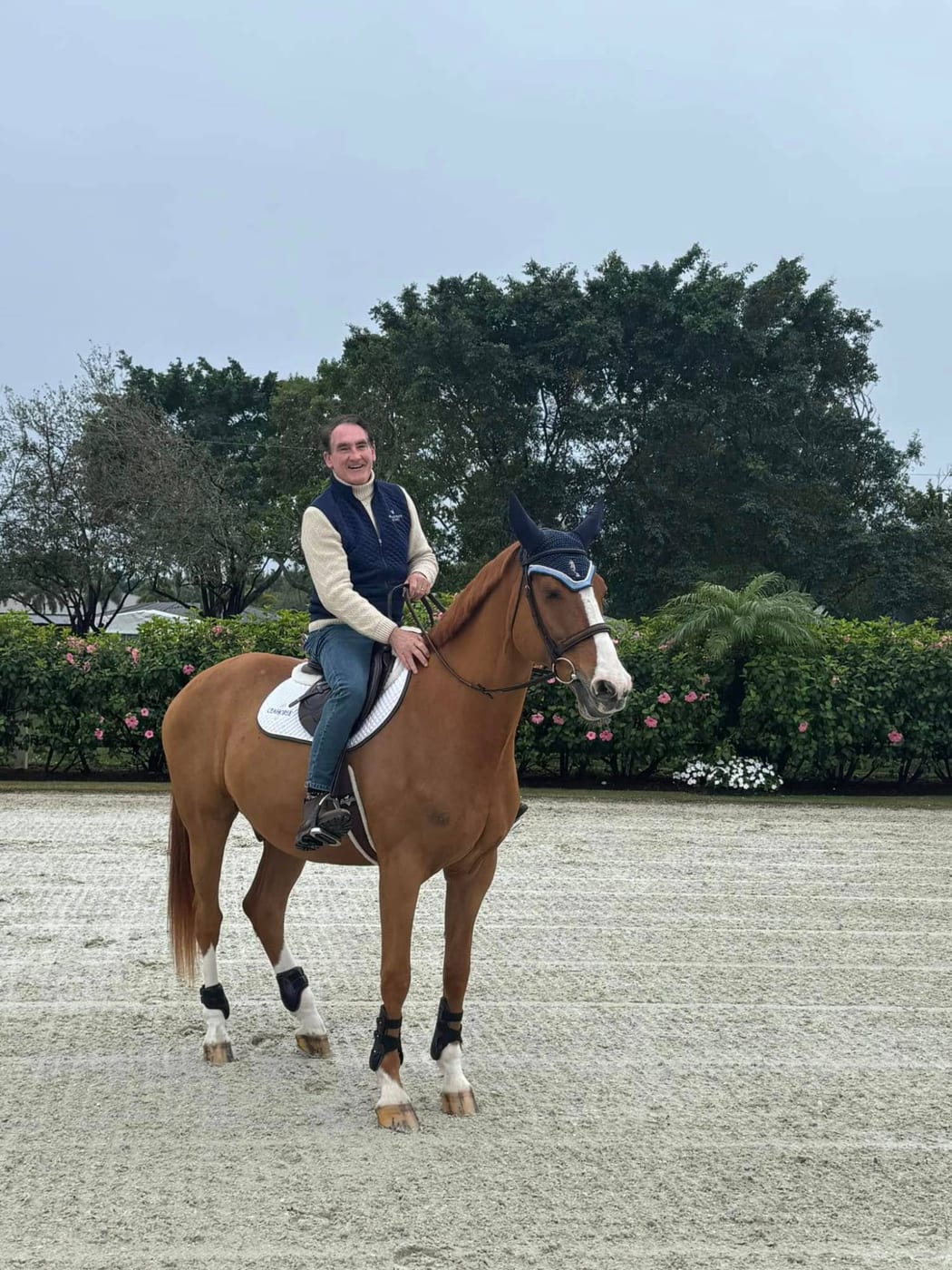 Bing Bush smiling in the saddle on a chestnut sport horse with a navy ear bonnet in a sand arena