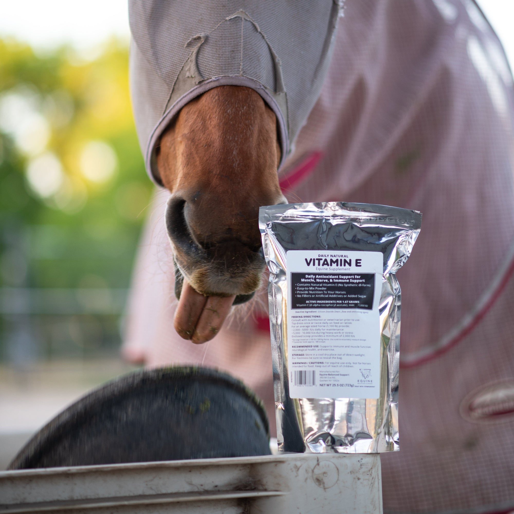 A bay horse in a fly mask reaching toward a pouch of Daily, Natural Vitamin E labeled for muscle, nerve, and immune support