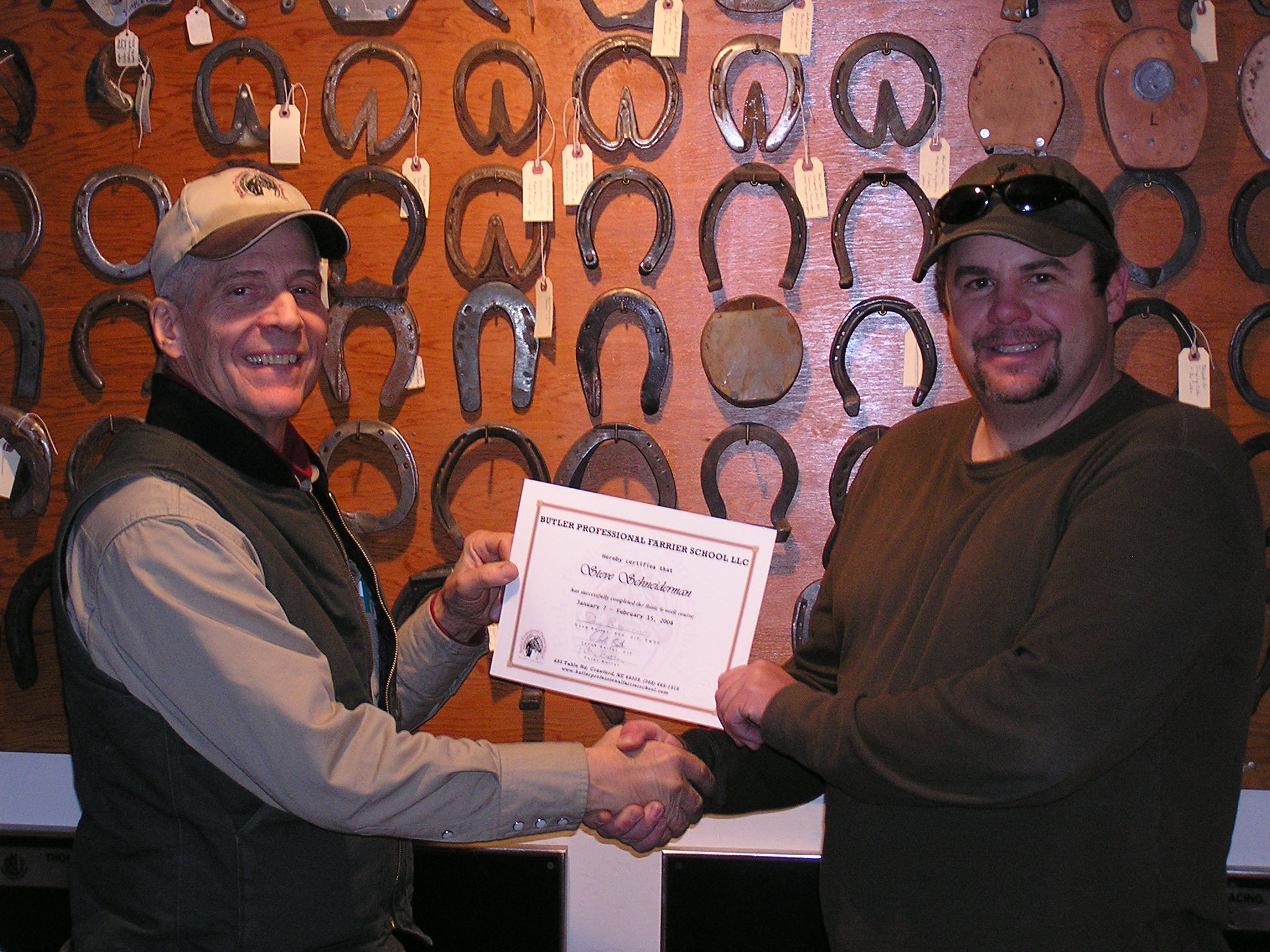 Dr. Doug Butler presenting a graduation certificate to a farrier student in front of a wall of handcrafted horseshoes