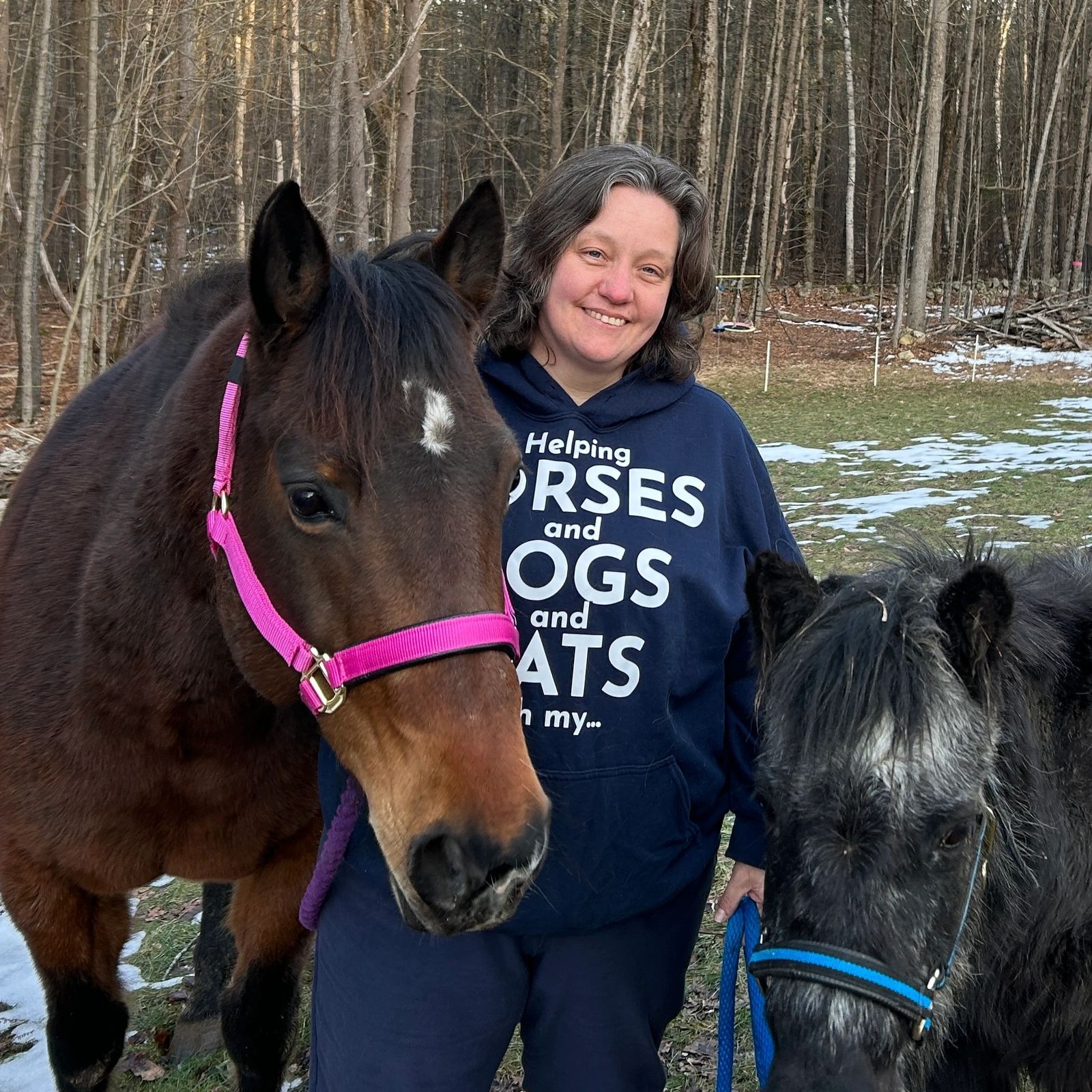 A smiling woman standing outdoors between a haltered bay horse and a dark pony