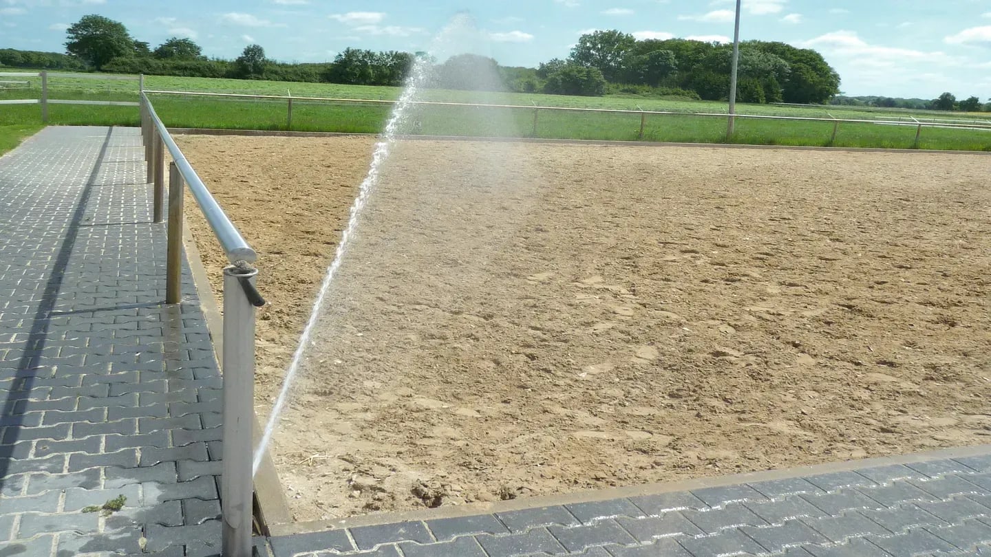 Corner-mounted sprinkler throwing a wide arc of water across an outdoor sand horse arena