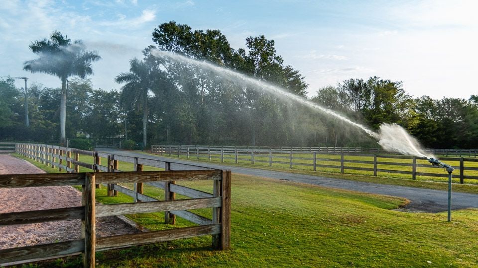 Large outdoor sprinkler throwing a long arc of water beside a white-fenced horse paddock in early morning light