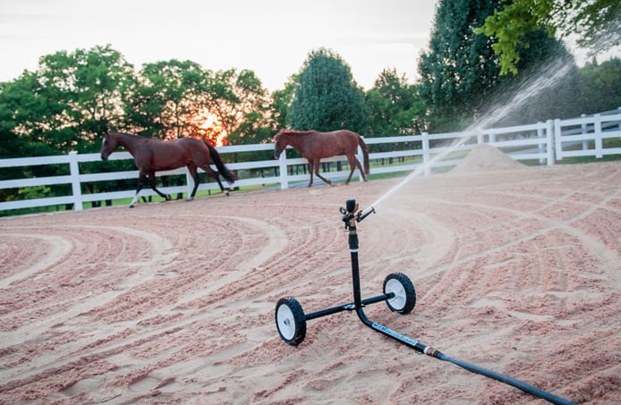 Travelling sprinkler watering a freshly groomed outdoor arena at sunset with two horses in the paddock behind