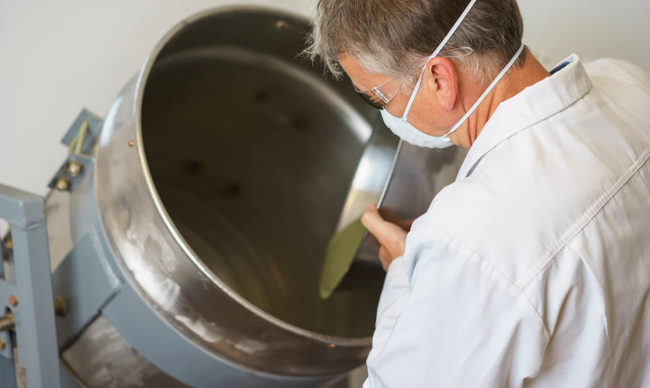 Sustenance Herbs maker in a lab coat and mask hand-mixing a batch of organic supplement in a stainless steel drum