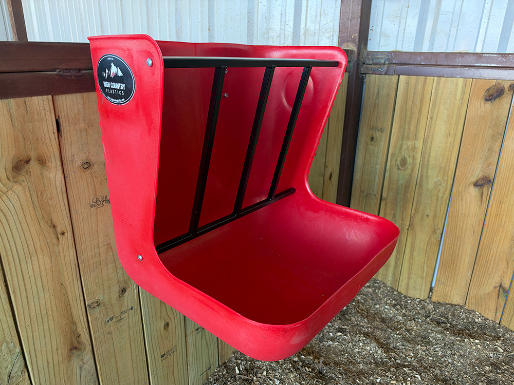 Red High Country Plastics hanging wall feeder with a black steel hay rack, mounted to a wooden stall wall above wood shavings