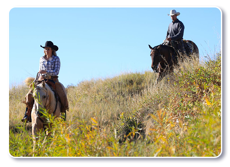 Two riders on horseback climbing a grassy, wildflower-dotted hillside on a working ranch