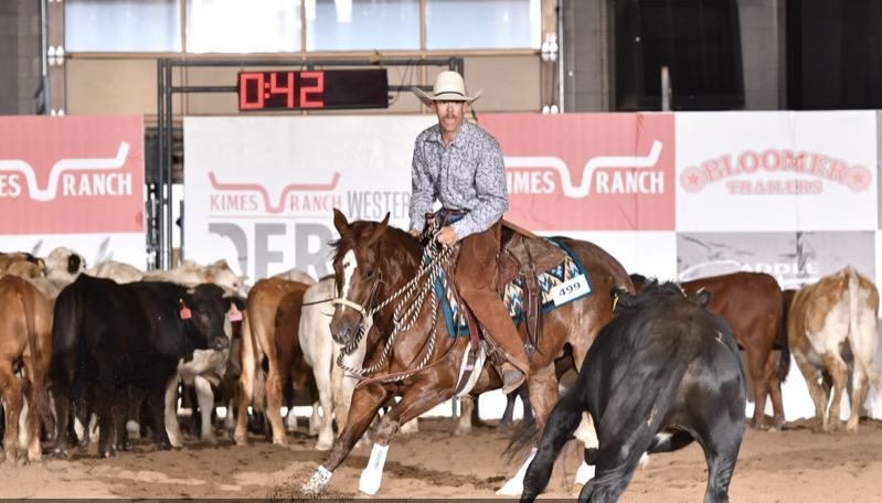 Lamar Community College horse program rider in cowboy hat and chaps riding a brown ranch horse with other horses behind