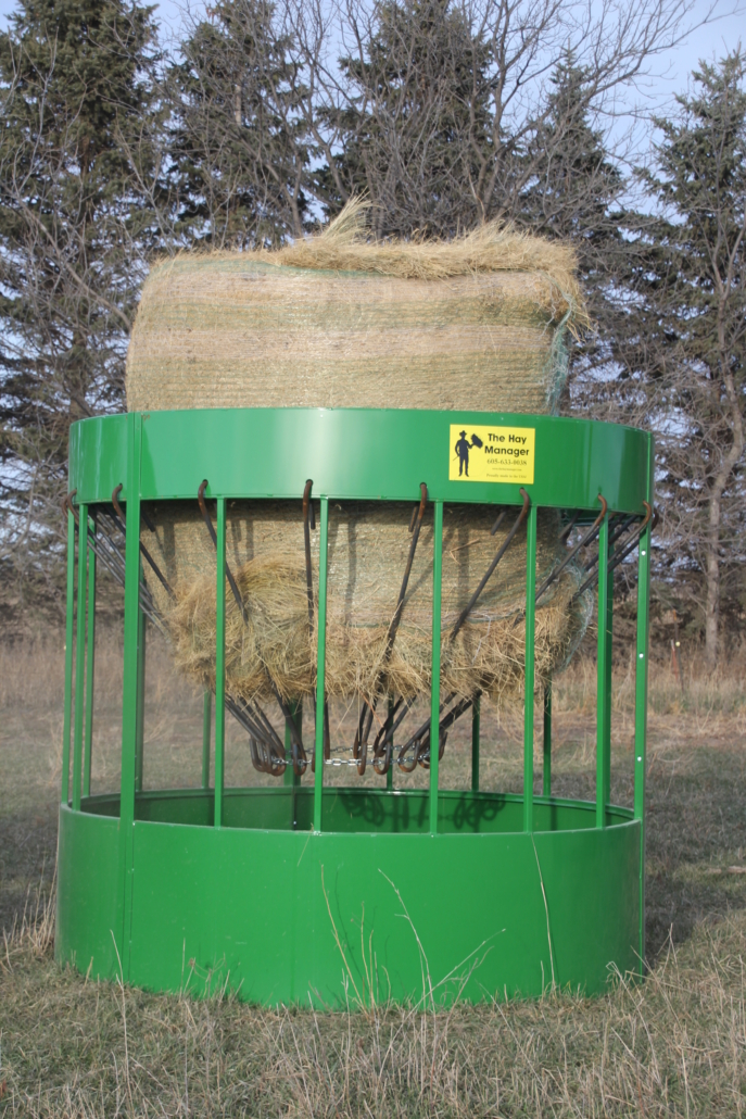 A green Hay Manager round bale feeder loaded with a full round bale of hay standing in a pasture, with the brand decal visible on the band